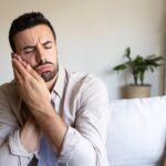 Caucasian man in pain suffering from toothache sitting on sofa at home. Copy space.