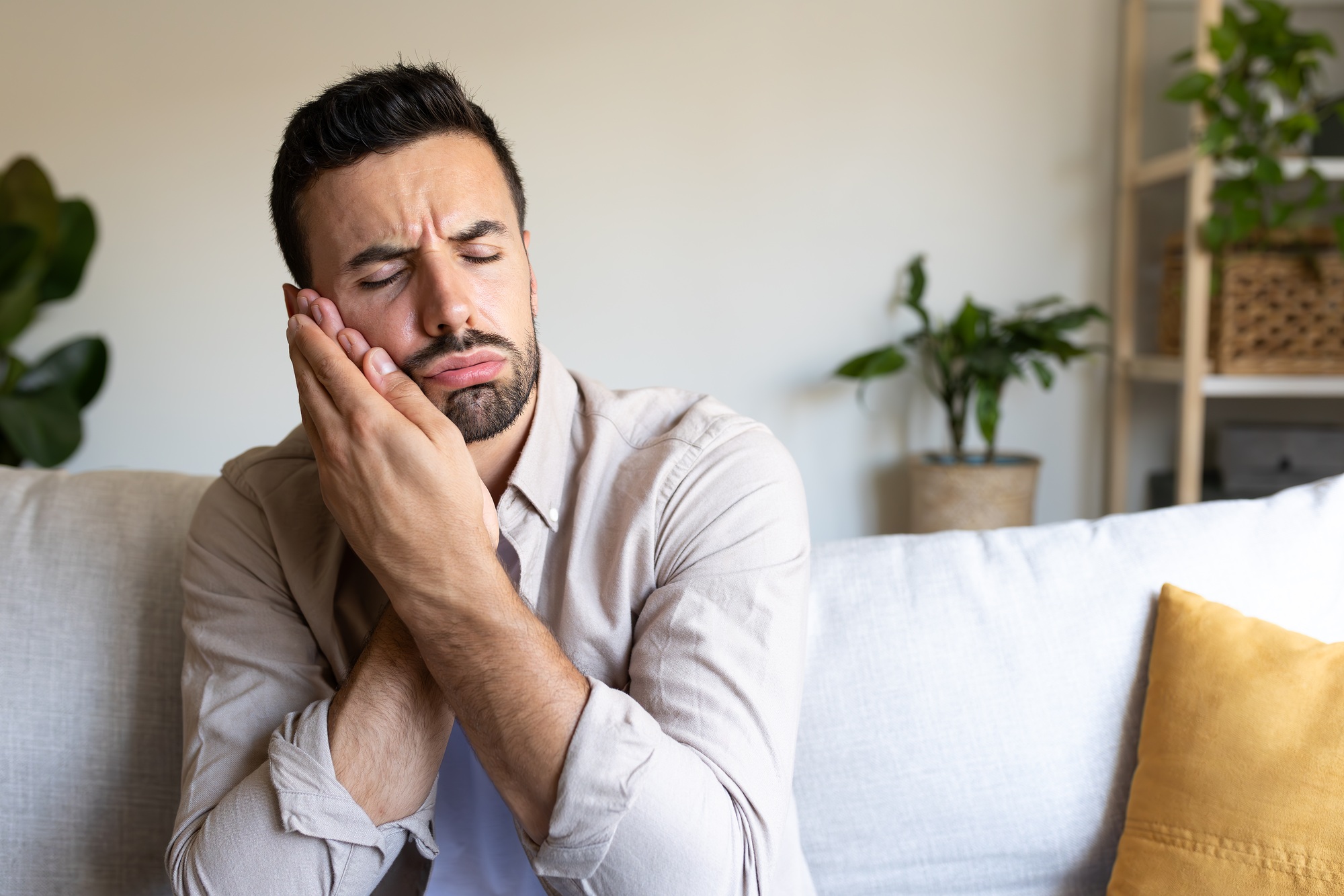 Caucasian man in pain suffering from toothache sitting on sofa at home. Copy space.